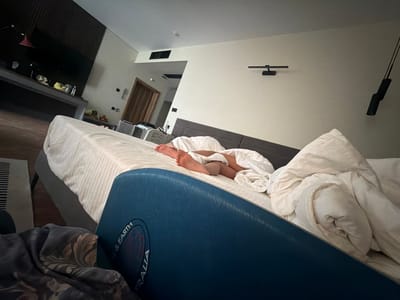The image depicts a bedroom scene. A person, likely a child, is sleeping on a bed covered with white linens. Their legs and feet are visible. In the foreground, a large blue object with text is partially visible, with the words "EARTH" and "TRALIA" discernible. To the left, a dark-patterned fabric is draped. In the background, a dark headboard is visible. Further back, there's a desk or console with items on it, including a lamp. A doorway is also visible in the background, suggesting another room or hallway. The walls are light-colored, and there are some light fixtures on the wall. This scene does not contain any elements that would indicate it is located in Morozovo, Russia.