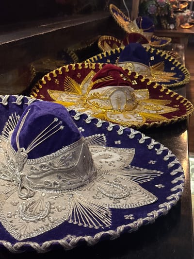 Four highly ornate traditional Mexican sombreros are displayed in an indoor setting, arranged diagonally from the foreground left to the background right on a dark, reflective surface.

The sombrero in the immediate foreground features a royal purple velvet brim and crown top, with a cream-colored main body and brim underside. It is extensively decorated with intricate silver embroidery, including stylized patterns, small sequins, and a silver corded trim along the brim edge. Four slender silver metallic elements protrude from the apex of the purple crown.

Behind this, a second sombrero presents a burgundy red velvet brim and crown top, with a cream-colored body. It is adorned with detailed gold embroidery, including sunburst patterns and cross motifs, and a gold corded trim. Similar metallic elements, in gold, are visible on its crown.

A third sombrero, positioned further back, exhibits a royal blue velvet brim and crown top with gold embroidery and trim, consistent in style with the burgundy sombrero. The fourth sombrero, partially obscured in the far background, also appears to have a blue brim with gold embroidery.

The sombreros rest on a dark, flat surface, likely a counter or shelf. Behind them, a dark, elevated horizontal surface is visible. The background is dark and blurred, with faint hints of other objects, possibly decorative elements or glassware, suggesting a commercial interior environment, such as a restaurant, bar, or shop. No individuals are present. The specific location within Atizapán de Zaragoza, Mexico, cannot be determined from the visual evidence provided.
