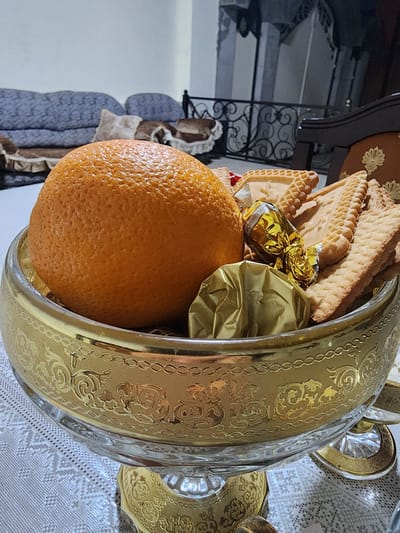 A close-up, eye-level, indoor shot features a golden, ornate glass bowl filled with an orange, several wrapped candies in gold foil, and an assortment of rectangular cookies. The bowl rests on a white, textured tablecloth. In the background, a blue sofa with patterned cushions is visible, along with furniture and architectural details that suggest an interior residential setting. The lighting is moderate, casting soft shadows. The overall impression is one of a decorative table setting. This image is not identifiable as being located in Gudermes, Russia.