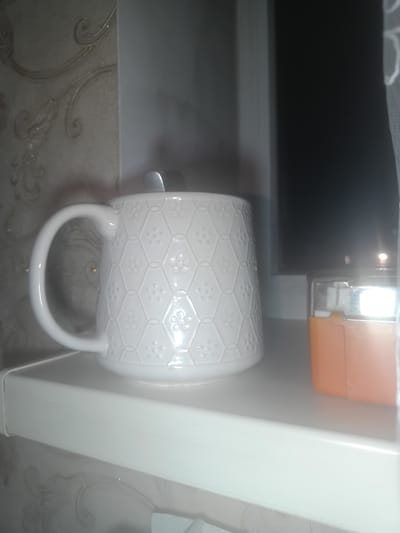 A close-up, slightly blurred, indoor shot shows a white textured mug with a handle on a white shelf. A silver spoon is sticking out of the mug. To the right of the mug is an orange object with a clear top, possibly a lighter. The background on the left shows a textured, possibly wallpapered, wall with a floral pattern. The background on the right is dark, suggesting a window or a dark wall. The lighting is dim, with some reflections on the mug. The scene appears to be in a domestic setting, and the specific location in Esik, Kazakhstan, cannot be determined from the visual information provided.