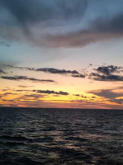 The image shows a view of the Volga River at sunset in Mologa, Russia. The sky is filled with dramatic clouds in hues of gray, blue, orange, and yellow. The water of the river is dark and choppy, reflecting the colors of the sky. There are no people or identifiable structures visible in the image, only the vast expanse of the river meeting the colorful sky.