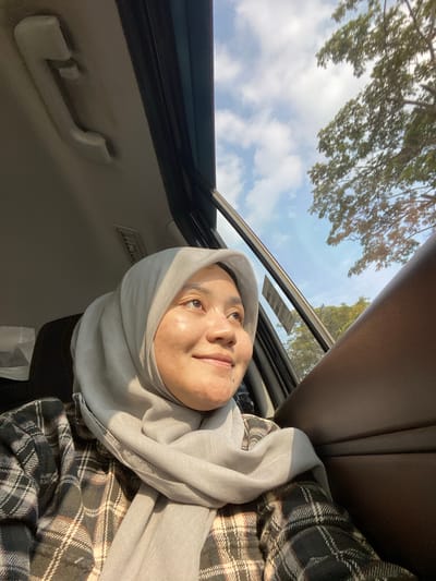 A young woman wearing a light gray hijab and a plaid shirt looks out of a car window. The woman is positioned in the foreground, with her face turned to the side, illuminated by sunlight. Her expression is serene and contemplative. Outside the car, a blue sky with scattered clouds is visible, along with the branches of a leafy tree. The interior of the car, including the door panel and a handle, are also present in the frame. The image was taken in Mandar Sari, Indonesia.