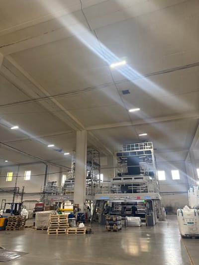The image depicts the interior of a factory, likely a plastics manufacturing facility, located in Osentsy, Russia. The space is vast, with a high ceiling supported by concrete columns. Natural light streams in through rectangular windows spaced along the far wall, and overhead fluorescent lighting illuminates the production floor.

Dominating the center of the scene is a large, multi-story industrial machine, likely a film extrusion or blowing machine, with complex scaffolding and control panels. Two individuals, presumably workers, are visible operating or attending to this machinery.

In the foreground and to the left, a yellow forklift is parked next to stacks of wooden pallets. Some pallets are laden with white sacks, possibly raw materials, and others are stacked empty. Additional pallets with various items are scattered across the floor, including what appear to be rolls of plastic film.

To the right, several large white sacks, likely containing industrial materials, are stacked on pallets. The floor is concrete, and there are power cables running overhead. The overall impression is one of an active industrial environment focused on production.