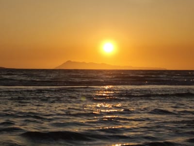 The image depicts a sunset over the sea, likely taken in Agios Stefanos, Greece, given the prompt's context. A large, bright sun is positioned centrally in the sky, which is a gradient of orange and yellow hues. A dark, silhouetted mountain range is visible on the horizon behind the sun. The sea is dark and choppy, reflecting the sunlight in streaks across the water's surface. There are no people or discernible objects in the foreground other than the water. The scene appears tranquil.