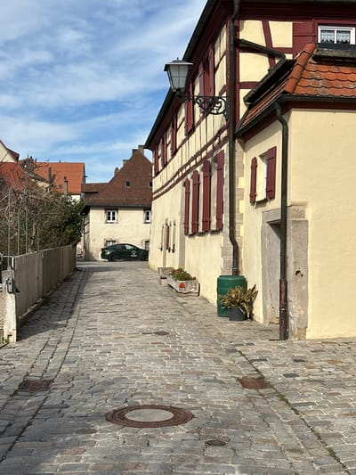This image depicts a narrow, unpopulated cobblestone alleyway in Weißenburg in Bayern, Germany, under a partly cloudy sky. The street surface is composed of irregular grey cobblestones, featuring multiple circular metal manhole covers and rectangular drain grates, and recedes uphill into the background.
On the right side, a prominent two-story half-timbered building with a cream-colored facade and dark red wooden beams features numerous windows, all fitted with closed red shutters. An ornate black metal street lamp with a glass lantern is affixed to the building's upper facade. Below, a concrete planter contains small plants, followed by a dark green plastic rain barrel, and a potted plant in a black pot. Adjacent to this, a cream-colored building with a terracotta tiled roof displays a dark brown rain gutter and downspout, and a stone-framed doorway.
On the left side of the alley, a light-colored wooden fence runs parallel to the street, behind which leafless trees and shrubs are visible. Further down the alley, a dark green Tesla Model S vehicle, featuring a circular white logo on its side, is parked facing away from the viewer. Additional traditional European buildings with terracotta tiled roofs and light facades are visible in the distant background. The sky overhead is blue with scattered white, wispy clouds. No individuals or active interactions are present in the scene.