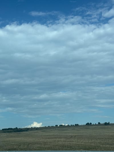 The image shows a vast, open landscape under a cloudy blue sky. The foreground is dominated by a dry, grassy field with subtle horizontal lines suggesting tilled soil or wind patterns. A metallic guardrail is visible at the very bottom of the frame, indicating a road or pathway. The horizon line is marked by a gently rolling hill with scattered trees and shrubs, their forms silhouetted against the sky. A large, fluffy white cloud formation is prominent over the hills in the middle distance, appearing almost like smoke. The sky itself is a mix of deep blue at the top, transitioning to lighter shades with numerous layers of wispy, greyish-blue clouds that obscure a clear view of the sun. The overall impression is one of a rural or natural setting, possibly during late summer or autumn given the dry vegetation. There are no people or specific man-made structures visible within the landscape itself, other than the guardrail. The image captures a wide expanse of sky and land, with the clouds being a significant feature of the scene. There is no information in the image to suggest a specific location within the city of Zheleznik, Bulgaria.
