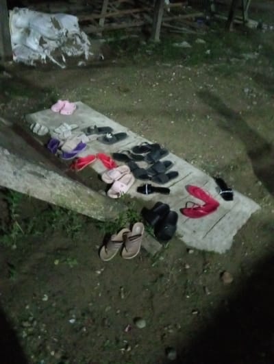 A collection of sandals and flip-flops are arranged on a concrete surface on the ground. The footwear varies in color, style, and material. The surrounding environment consists of grass and dirt. There is a pile of white sacks and a wooden structure in the background. The location is Pulorayeuk, Indonesia.
