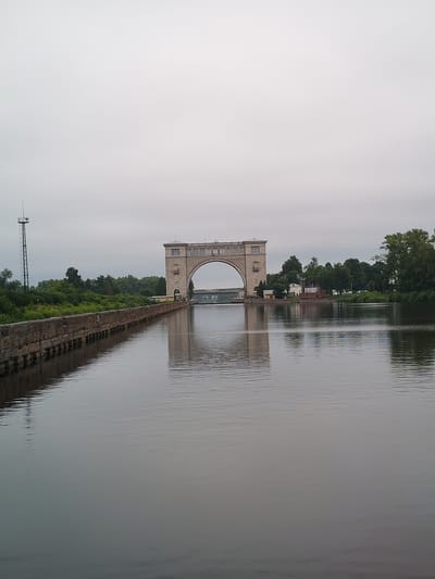 The image shows a large, arched structure over a body of water, likely a canal or river, in Faliukovo, Russia. The structure appears to be a lock or aqueduct, made of light-colored stone, with windows and decorative elements. In the foreground, the water is calm and reflects the overcast sky and the surrounding environment. To the left of the structure, there is a stone embankment with vegetation growing on top, and a tall metal tower with antennas. The right side of the image shows trees and some smaller buildings, possibly part of the lock's infrastructure. The overall scene is tranquil, with no visible people or activity.