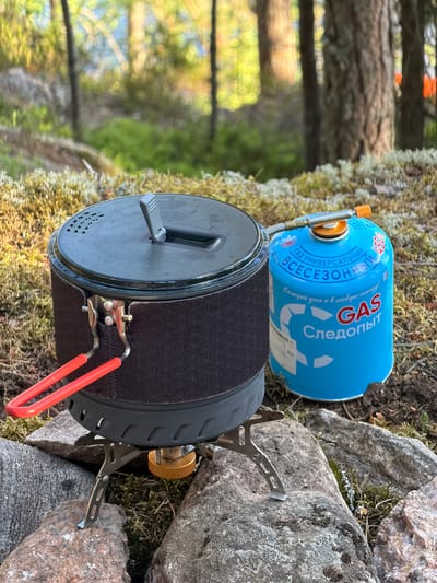 A camping stove with a blue gas canister sits on mossy ground and rocks, surrounded by trees in a forest. The stove is equipped with a pot with red handles and a black lid. The gas canister is labeled "GAS Следопыт" and "ВСЕСЕЗОННЫЙ." The scene appears to be in a natural, outdoor environment, possibly in the city of Ruch'i, Russia, given the presence of coniferous trees and moss.