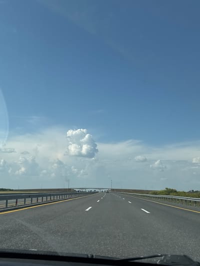 The image shows a view from a vehicle on a highway, likely in or near Алтынды ауылдық округі, Kazakhstan. The highway stretches forward under a bright blue sky with scattered cumulus clouds. The road is asphalt, marked with white lane lines and a yellow line on the right shoulder. Guardrails line both sides of the road. In the distance, a bridge or overpass is visible, with a few vehicles on it. The landscape on either side of the highway appears to be flat and somewhat barren, with some low vegetation. The focus is on the road and the expansive sky, suggesting a sense of travel and open space.