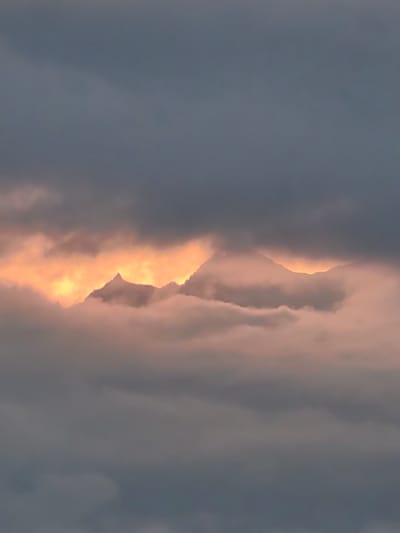 A photograph captures a dramatic sky over the city of Aleo, India. The scene is dominated by dark, moody clouds that obscure much of the sky, creating a sense of an approaching storm or the fading light of dusk. Peeking through a break in the clouds, the rugged peaks of mountains are illuminated by a vibrant, fiery orange glow, suggesting the setting sun or a powerful light source behind them. This glowing aperture in the clouds creates a striking contrast with the somber tones of the surrounding cloud cover. The mountains themselves appear silhouetted against this intense light, their jagged edges sharply defined. Wisps of lighter, pink-tinged clouds drift in front of and around the mountains, softening their appearance and adding depth to the scene. The overall impression is one of natural grandeur and the awe-inspiring power of the elements.