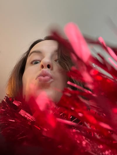 A single female subject is depicted in a close-up, low-angle indoor portrait. Her face occupies the upper-middle portion of the frame, with her eyes wide open and directed upwards. Her lips are puckered in a pronounced "kissing" gesture. Dark brown hair frames her face. A substantial amount of vibrant red, reflective tinsel-like material is prominently positioned in the foreground, extending across the bottom and up the right side of the image, partially obscuring the subject's neck and right side of her face. Portions of the tinsel appear blurred due to depth of field. The background consists of a plain, light-colored surface, consistent with an interior setting. The specific location in the city of Callbach, Germany, is unidentifiable from the visual evidence.