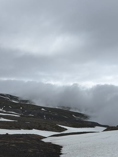 The image depicts a desolate, mountainous landscape under an overcast sky. The terrain is a mixture of dark volcanic rock and patches of snow, suggesting a high-altitude or cold environment. The slopes are uneven, with some areas appearing barren and others sparsely covered with low-lying vegetation. A thick band of clouds or fog rolls across the middle ground, obscuring the upper portions of the mountains and adding a sense of mystery to the scene. The foreground shows a clear expanse of snow, contrasting with the dark soil. There are no people or structures visible, emphasizing the wild and remote nature of the location.