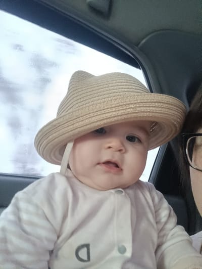 A baby wearing a light-beige, woven straw hat with two pointed 'ears' and a chin strap is positioned inside a vehicle in Simakovo, Russia. The baby has light skin, dark eyes, and a slightly open mouth, looking towards the camera. It is dressed in a light-colored, long-sleeved garment with horizontal stripes on the sleeves, a front snap closure, and a dark grey letter "D" printed on the chest. A portion of an adult's face, including dark-framed eyeglasses and dark hair, is visible on the far right, adjacent to the baby. The background shows a vehicle interior with a grey headliner and a window revealing a blurry, light exterior.