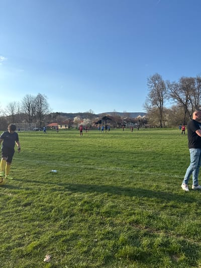 The image depicts a soccer field in Dushevo, Bulgaria. A soccer game is in progress with players in blue and red jerseys. The field is green with white lines. Bare trees surround the field. Two people are visible in the foreground. One person is fully in the frame at the bottom, wearing dark clothing and yellow socks. Another is partially visible in the top right, wearing a black shirt and jeans. Buildings and a mountain are in the background. A blue sky is overhead with contrails. A water bottle is visible on the field.