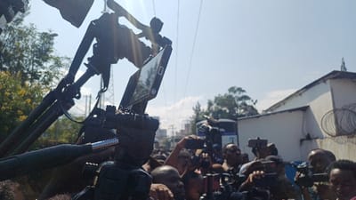 This image depicts a crowded outdoor scene in Kabinzi, Rwanda, under a clear, bright blue sky with visible sunlight causing lens flare. The perspective is low-angle, looking upwards and through a dense gathering of individuals.

In the immediate foreground, a professional video tripod with an attached smartphone is prominently featured, positioned slightly to the right of the center. The smartphone, dark-colored with visible side buttons, is mounted horizontally, its screen reflecting light. Behind and around this setup, numerous individuals are visible, primarily from the chest up or as disembodied heads. Many are holding up various recording devices, including other smartphones (some mounted on selfie sticks or small tripods) and professional video cameras with attached microphones or accessories. The crowd appears to be intently focused on an unseen subject, with several arms raised, holding devices to capture footage.

In the midground and background, an urban or semi-urban environment is present. To the left, green foliage from trees is visible, with hints of yellow flowering plants. Overhead, multiple thin power lines or communication cables stretch across the sky. To the right, a white building with a dark roofline is present, featuring what appears to be razor wire coiled along a wall or fence. Farther back, additional buildings and more distant green trees are discernible under the bright sunlight. The overall impression is a media-heavy event or public gathering.