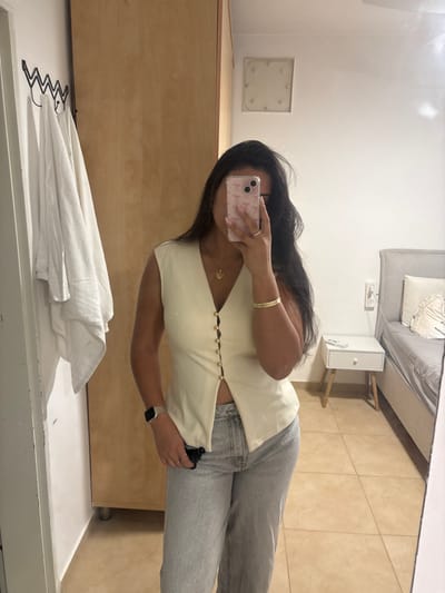 A female individual is depicted in a mirror selfie within an indoor room, identified as being in Maale Adumim, Palestinian Territory. The subject has long dark hair and is dressed in a cream-colored sleeveless V-neck top with gold-toned buttons down the front, unbuttoned to expose the midriff, paired with light-wash denim jeans. She holds a light-colored smartphone with a clear case in her right hand, obscuring her face. A gold heart pendant necklace is visible, along with a gold-colored bracelet on her right wrist, a silver-toned smartwatch on her left wrist, and a silver ring on her right hand's ring finger. A dark object is partially visible in her left hand, tucked into her jeans pocket.
The room features light brown square-tiled flooring and white walls. To the left, a light wooden two-door wardrobe stands adjacent to a white door. A black wavy metal wall hook holds a folded white towel to the left of the wardrobe. To the right, a bed with a light grey upholstered headboard, grey bedding, and a decorative white textured pillow is visible. A white nightstand with a single drawer and light wooden tapered legs is positioned next to the bed. On the upper right wall, a square light fixture or vent cover with eight visible screws is present.