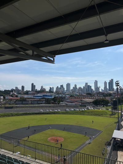The image displays an elevated view of a baseball field in Curundú, Panama, overlooking a densely developed urban landscape under a clear, partially cloudy daytime sky. The foreground features the underside of a stadium roof, composed of dark gray I-beams and white ceiling panels, with green stadium seating and a black metal railing visible at the bottom. The central element is a green grass baseball field with a dark gray dirt infield, a reddish-brown pitcher's mound, and a home plate area. Approximately 10-15 individuals, dressed in athletic attire, are present on the field, positioned as if engaged in a baseball game or practice; several are in the infield and outfield, one is near the pitcher's mound, and another appears to be at home plate. A dugout structure on the right side of the field houses additional figures. Beyond the field, a paved parking lot contains numerous white cars. The mid-ground comprises a mix of lower-rise commercial and industrial buildings. The background is dominated by a panoramic view of Panama City's skyline, characterized by a dense cluster of numerous modern high-rise buildings and skyscrapers.