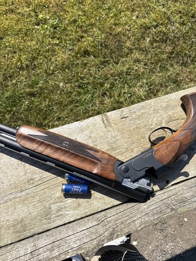 An over-under shotgun, identified as an ATA Arms model, rests horizontally on a weathered wooden surface under bright sunlight at an outdoor location in Wold Newton, United Kingdom. The shotgun, featuring blued barrels and receiver with a gold-colored trigger, displays a dark brown wooden stock and fore-end with checkered grips, including a stylized double-cross logo on the fore-end. Its action is open, exposing the breech. Two blue shotgun shells, labeled "HULL" and "SUPER FAST FIBRE," lie near the barrels on the wooden surface. The immediate background shows green grass. The lower right foreground reveals a partial view of a person's lower leg and foot, wearing a light-colored sneaker and dark trousers or gaiters, positioned in front of the wooden surface.
