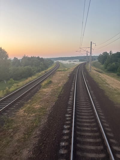 The image shows a view from a moving train on a railway track in an outdoor rural environment. The sky is a gradient of light blue and pale orange, indicating either dawn or dusk. Two sets of railway tracks run parallel, curving into the distance. The tracks are made of gravel and sleepers, with overhead electrical wires supported by pylons. The surrounding landscape is lush with green trees and vegetation, with a light mist or fog visible in the distance over a field. The scene appears to be set in the region of Пратасевіцкі сельскі Савет, Belarus, given the characteristic railway infrastructure and natural landscape of the area. There are no people or specific buildings visible in the immediate vicinity.
