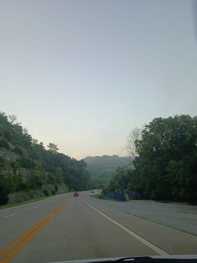 This media file depicts a scenic view from a moving vehicle on a highway. The scene is dominated by a road that curves into the distance, bordered by lush green trees and a rocky hillside. Two yellow lines are painted on the road, indicating a highway. Several cars are visible on the road, suggesting that it is a public thoroughfare. In the background, the landscape is hilly and covered in dense vegetation. The sky is a light, hazy blue, which could indicate early morning or late afternoon, or perhaps a slightly overcast day. There are no people clearly visible in the image, but the presence of cars implies human activity. A sign with text is visible on the left side of the road, but the text is too small to be legible. The overall impression is one of a serene journey through a natural landscape.