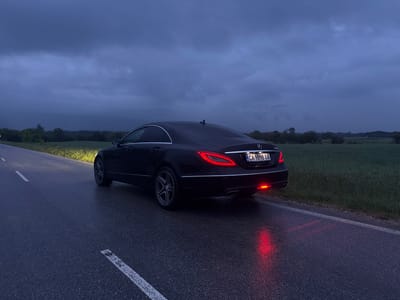 The image depicts a black Mercedes-Benz CLS parked on a wet asphalt road. The car's taillights and left reverse light are illuminated. The license plate reads "CA 9898 XA." The road has a white line marking its edge. Adjacent to the road is a field with green vegetation. In the background, a dark, overcast sky dominates the scene, with hints of trees and distant hills. The location is inferred to be near Kliment, Bulgaria, based on the license plate code.