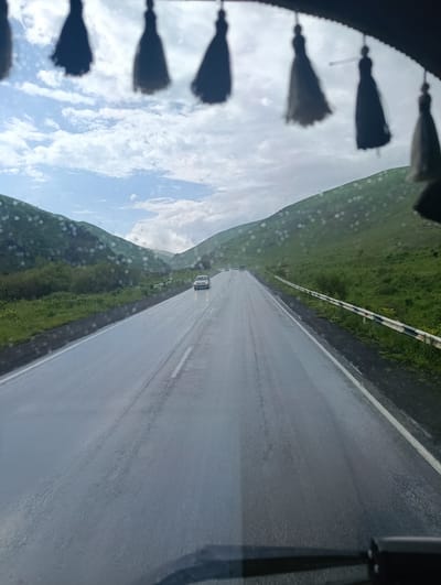 This media file depicts a view from inside a vehicle traveling on a wet, asphalt road. The road stretches ahead, flanked by green, rolling hills that create a valley-like setting. The sky is partly cloudy with dramatic, white clouds against a blue backdrop. Several dark, tassel-like ornaments hang from the top of the frame, obscuring a portion of the sky and the upper edges of the hills.

Several vehicles are visible on the road. In the distance, on the right side of the road, a single white car is driving. Further ahead, another car appears to be in the distance. The road surface is wet and reflective, suggesting recent rain or damp conditions. Small water droplets are visible on the windshield from which the photo is taken, further reinforcing the idea of wet weather.

The landscape appears to be rural, with lush green vegetation covering the hillsides. There are also some areas of denser bush or small trees along the side of the road. A white and black striped guardrail runs along the right side of the road, and a yellow triangular road sign with a red border is visible next to it. In the far distance, a tall structure, possibly a power pylon, can be seen.

The time of day appears to be daytime, likely late morning or afternoon, given the brightness of the sky. The overall mood is somewhat somber due to the wet road and overcast skies, but the vibrant green of the hills offers a sense of natural beauty. The presence of the tassels suggests the photo was taken from the interior of a car or truck, possibly a passenger vehicle adorned with such decorations. No visible text is present in the image. The location is Semyonovka, Armenia.