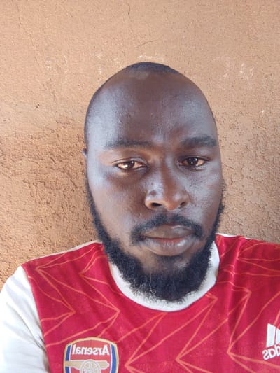 A close-up photograph depicts a dark-skinned man with a bald head and a beard. He is wearing a red and white soccer jersey with a logo that is partially visible, appearing to be an Adidas logo. The background is a textured, light brown wall. The setting appears to be indoors during the day, as determined by the lighting on the man's face and the wall behind him. The photograph does not include sufficient information to verify the location in Mulke Barikin Sayejo, Nigeria. There are no discernible actions or interactions taking place in the image.