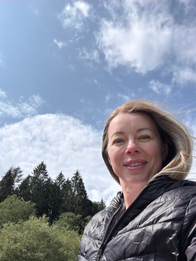 A woman with blonde hair and blue eyes is taking a selfie outdoors. She is wearing a black puffer jacket. The background shows a blue sky with white clouds and a line of green trees. The image is a close-up, with the woman's face and shoulders filling most of the frame.