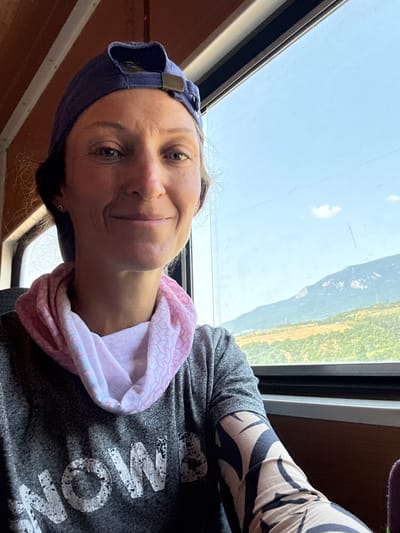 A medium shot selfie shows a woman wearing a blue baseball cap, a pink and white patterned bandana around her neck, and a gray t-shirt with white lettering that reads "NOW." She is smiling and looking at the camera. Her right arm is visible, covered in a tan sleeve with a black abstract pattern. She is seated next to a window, with a blurred view of mountains and a valley outside. The interior of what appears to be a train car is visible behind her, with brown wooden paneling and white trim. The image is taken in daylight.