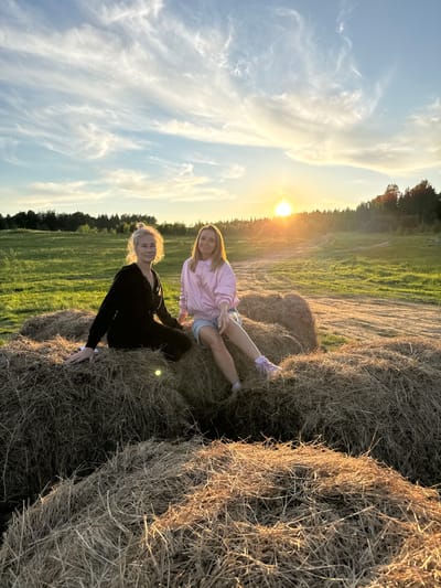 Two women are seated on large hay bales in a golden-hour field, bathed in the warm light of a setting sun. The image captures a serene outdoor scene, likely in a rural or agricultural area near Стеклянный, Russia.

One woman, positioned in the foreground to the left, has blonde hair tied back and is wearing a black long-sleeved top and dark pants. She is smiling gently at the camera. Behind and to her right, a second woman with long blonde hair is dressed in a pink long-sleeved top and denim shorts, also smiling towards the viewer. Both appear relaxed and joyful.

The hay bales they sit upon are textured and appear to be made of dried grass or straw, strongly illuminated by the low sun. Beyond the hay bales, a vast green field stretches into the distance, bordered by a line of trees on the horizon. The sky is a prominent feature, displaying a bright, setting sun on the right, which casts a golden glow and produces lens flare. The sky transitions from a soft blue at the top to vibrant oranges and yellows near the sun, with scattered wispy clouds. The overall ambiance is peaceful and pleasant. No text is visible in the image.