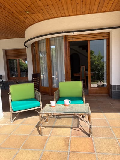 The image depicts an outdoor patio area with tiled flooring in Can Ribas, Spain. Two wicker chairs with bright green cushions and teal seat cushions are arranged facing a small, glass-topped coffee table. On the table, two mugs with orange rims are present. Behind the chairs, a set of sliding glass doors opens into an interior space, revealing a dining table and chairs. The exterior of the building features a curved wooden ceiling and white stucco walls. Sunlight casts shadows on the patio, indicating a daytime scene.