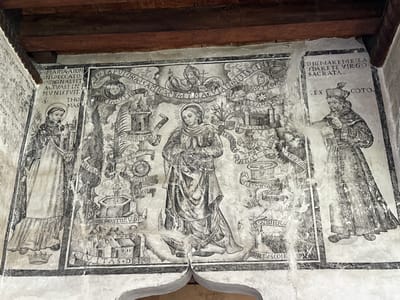 This is a black and white fresco painted on a wall within the Church of San Miguel Arcángel in Huejotzingo, Mexico. The fresco depicts a religious scene with multiple figures and symbolic elements. In the center is a female figure, likely a saint, with her hands clasped in prayer and a halo above her head. She is dressed in robes. To her left stands a male figure in monastic attire, also with a halo, holding a staff and looking towards the central figure. To the right of the central figure is another male figure, also in religious garb and wearing a distinctive hat, holding a book or scroll and appearing to read.

The fresco is surrounded by elaborate scrolls and banners containing Latin inscriptions, though they are difficult to read in detail due to the age and condition of the painting. Various architectural elements, such as towers and cityscapes, are depicted, along with symbolic objects like crowns, stars, and plants. The overall composition is complex and laden with religious symbolism. The fresco shows signs of age and wear, with some areas appearing faded or damaged. The background is a textured, light-colored wall, and the wooden ceiling beams are visible at the top of the image.