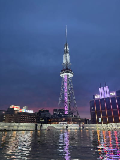 The image captures the Nagoya TV Tower and the surrounding cityscape at dusk, with a hint of pink and purple in the sky. The TV tower, a prominent landmark in Nagoya, Japan, stands tall with its lattice structure illuminated by purple lights along its central section. To the right of the tower is a modern building with "ESTHE" and "TWIN" displayed in illuminated lettering. In the foreground, a body of water reflects the lights of the buildings and the sky, with several people standing along a white barrier overlooking the water. The water's surface is rippled, creating distorted reflections of the urban landscape. Several smaller buildings with illuminated signs are visible in the background, contributing to the vibrant urban setting.