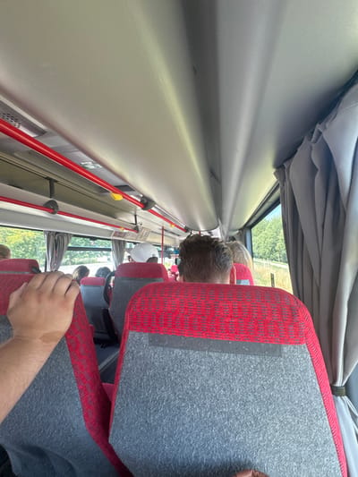 The image shows the interior of a bus in Wolfsthal, Austria. The perspective is from a passenger's seat, looking forward and slightly to the right. The seats are upholstered in red and grey fabric with a diamond pattern. Red grab rails run along the ceiling of the bus. Through the windows, trees and greenery are visible, suggesting a rural or semi-rural setting. A hand is resting on the back of a seat in the foreground. Several passengers are visible, their backs to the camera, some wearing hats. The overall impression is of a journey in progress.
