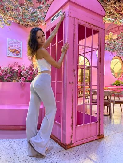 A woman with long, wavy dark hair stands next to a vibrant pink, vintage-style telephone booth. She is wearing a white bandeau top with a pink trim and loose, light blue flared pants. Her left hand rests on the top of the telephone booth, and her right hand is on the booth's side panel. The background is also predominantly pink, with floral decorations hanging from the ceiling and a large arrangement of pink roses to the left of the woman. A framed picture displaying what appears to be fried chicken is visible on the pink wall behind the roses. In the background, through the glass of the telephone booth, a cafe interior is discernible, with tables and chairs and circular mirrored decorations on the wall. The floor is tiled with a speckled gray and white pattern. This scene is located in a cafe in Tiến Thành, Vietnam.