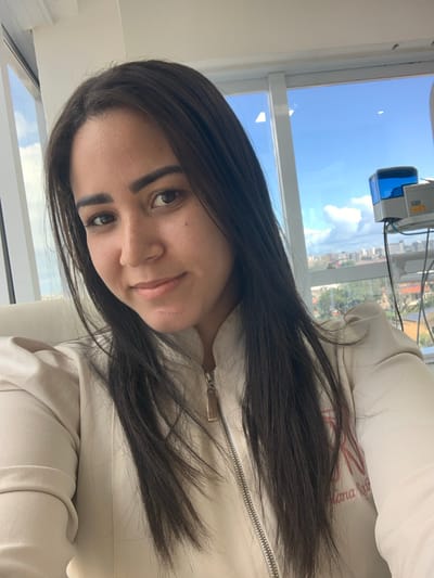 The image displays a female individual, appearing as an adult, positioned indoors for a selfie. She has long, straight dark hair, brown eyes, and light skin with freckles visible across her nose. She is wearing a light beige or off-white uniform jacket featuring a silver front zipper. An embroidered logo, "RN" with additional text "Eliana Ratk...", in a dark pink or red hue, is visible on the jacket's right chest area. Her expression is a subtle smile, and she looks directly at the camera.
The background indicates an indoor setting with expansive windows offering a view of an urban landscape in the city of Lauro de Freitas, Brazil. The exterior scene features a clear blue sky with scattered white clouds above a collection of buildings and green vegetation. Through the window on the right, a medical or laboratory device, characterized by a blue top and silver-grey base with visible wiring, is present. A light-colored chair or furniture piece is partially discernible behind the individual's left shoulder. No other individuals or direct interactions are depicted in the scene.