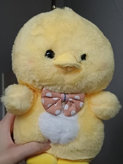 A close-up, vertical image depicts a yellow, fluffy chick plush toy held by a human hand. The plush features two small black bead eyes, a short orange fabric beak, and a prominent white oval patch on its chest. A pink fabric bow tie with white polka dots is secured around its neck. A small metal ring is visible at the apex of the plush's head. A light-skinned hand, with the thumb and fingers visible from the lower-left, supports the toy. The background is a plain, dark gray surface, out of focus. This scene is located in 서대문구, South Korea.