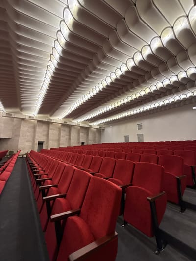 The image shows the interior of an auditorium or theater with two distinct sides. On the left, a wall features a textured, scalloped pattern illuminated by integrated lighting, creating a rhythmic play of light and shadow. This side appears to be a decorative architectural element. On the right, rows of vibrant red theater seats are visible, receding into the distance. The seats are upholstered and have dark metal armrests and supports. The perspective is from a high vantage point, looking down and across the space. The overall lighting suggests an indoor setting, possibly during an event or before one. The architecture, with its distinct treatment of the walls and the rows of seating, points to a venue designed for performances or presentations. No people are visible in the image. The backdrop is an empty auditorium, suggesting anticipation or a moment between events.