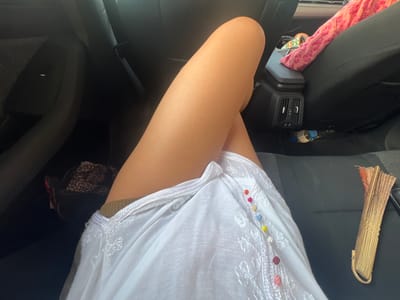 A person's legs are crossed, with the left leg on top of the right. They are wearing a white dress with embroidered details and colorful beads. The person is sitting in the back seat of a car, which appears to be a dark interior. A patterned bag and a pink scarf are visible on the seat behind them. On the floor of the car, a water bottle and a fan are present. The environment is indoors, within a vehicle. This image was taken in Felgueiras, Portugal.