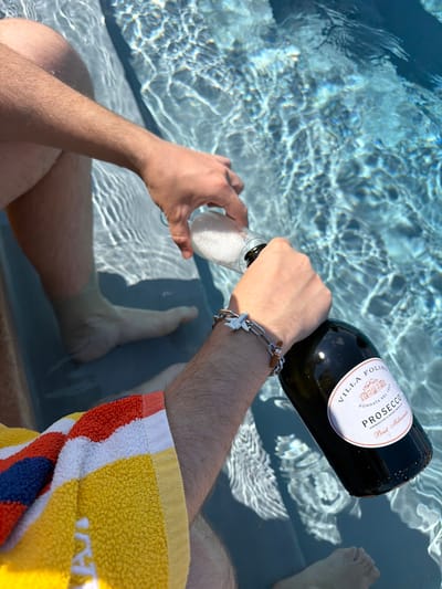 In this high-angle shot, a person is relaxing in a swimming pool, pouring Prosecco into a glass. The water is clear and shows ripples and reflections from the sunlight, suggesting it is daytime and likely sunny. The setting appears to be a pool, possibly outdoors, given the natural light. The person's legs and feet are submerged in the water, and a brightly colored towel, featuring stripes of yellow, white, orange, and blue, is draped over their arm. The focus is on the action of pouring the sparkling wine.

Visible text on the bottle label reads "VILLA FOLINI Prosecco". The bottle itself is dark glass, and the label is white with red and black text, and a crest. The person is wearing a silver bracelet with a charm shaped like an airplane. The overall mood conveyed is one of leisure and enjoyment. There are no other people visible in the frame. The context provided places this scene in Asciano, Italy.