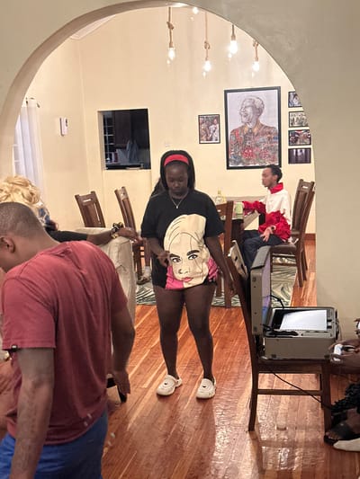 A gathering indoors shows a group of people. A woman in a black t-shirt with a face graphic and a red headband poses centrally. Behind her, a person in a red and white outfit leans over a table holding what appears to be a projector. Other individuals sit or stand around, one wearing a red shirt and blue shorts. Artwork hangs on the walls, and the lighting includes hanging bulb fixtures. The scene is warm with natural wood flooring.