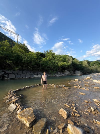 The image depicts a person standing in a shallow river on a sunny day. The person, a young male with fair skin and short brown hair, is wearing black swim trunks. He is positioned in the middle of the river, with his legs submerged in the water up to his knees. The riverbed is composed of rocks and pebbles of various sizes, with some larger stones visible along the bank.
The setting is a natural landscape with lush green trees lining the riverbank on the left side of the image. The trees appear dense and healthy, suggesting a temperate climate. In the background, a mountain slope covered in greenery can be seen. The sky above is a vibrant blue with scattered white clouds, indicative of fair weather. A suspension bridge-like structure with metal cables is visible on the left, partially obscured by trees, suggesting human infrastructure in the vicinity.
The lighting suggests it is daytime, likely late afternoon due to the warm tones of the sunlight reflecting off the water and rocks. The overall mood conveyed is one of relaxation and enjoyment of nature. There is no visible text in the image. The location context is Krasnoye, Russia.