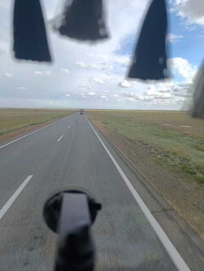 The image shows a view from a vehicle moving along a straight, paved road that cuts through a vast, flat, grassy plain. The sky is partly cloudy with a mix of white and gray clouds against a blue background. In the distance, a white truck is visible driving on the same road. The foreground is slightly blurred, showing the edge of the road and the roadside terrain. Dark, out-of-focus shapes are visible at the very top of the frame, possibly representing parts of the vehicle's interior or exterior. The overall scene depicts a journey through a desolate, open landscape, likely in a rural area. The location is identified as Қалыбай, Kazakhstan.