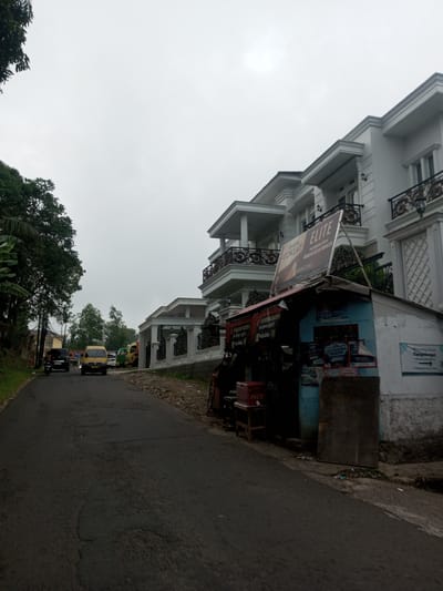 This image depicts a street scene in Houaythat, Laos, under an overcast sky. The foreground features a dark asphalt road with visible cracks and patches, extending towards the left into the distance. On the left side of the road, a steep embankment is densely covered with green foliage, including large trees. Several vehicles are present on the road further away: a dark-colored sedan, a yellow van, and a brightly colored truck, along with a partially visible motorcycle. On the right side of the road, a small, single-story, makeshift shop with a pitched roof stands prominently. The shop displays various advertisements, including "DJARUM SUPER," "MAGNUM FILTER," and a large billboard for "COKLAT ELITE" above it, all with Indonesian text. A red Coca-Cola cooler and a wooden stool are visible outside the shop. Behind the shop, a multi-story white building with a classical architectural style features ornate black and gold balustrades on its balconies, columns, and decorative wall elements. A white fence with decorative black accents surrounds the property of this building. No individuals are distinctly visible in the scene.