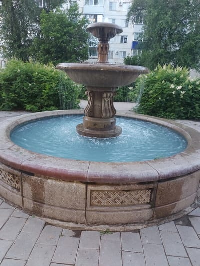 A stone fountain is centrally located in an outdoor urban setting. The fountain is tiered, with water cascading from the top basin into a larger lower basin filled with blue water. The fountain's base is surrounded by a paved walkway made of light-colored square tiles. Lush green bushes flank the fountain, and in the background, a multi-story apartment building with white and grey siding is visible. Trees with dense green foliage are interspersed between the bushes and the building. The overall environment suggests a residential area in Chapaevsk, Russia, with the fountain serving as a public amenity. There are no people visible in the image, and the fountain appears to be in operation.
