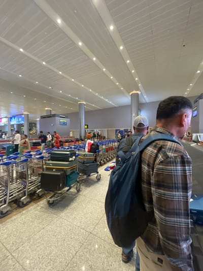 The image depicts the interior of an airport in Gollapalle Khurd, India, specifically in the baggage claim area. A number of people are seen standing around baggage carousels, some with luggage carts. The man in the foreground is wearing a brown plaid shirt and a blue backpack, facing away from the camera. The ceiling is a modern, modular design with integrated lighting. The floor is tiled with a light-colored, speckled pattern. In the background, a digital display screen shows flight information. The overall atmosphere appears to be one of transit and waiting, common in airport environments.