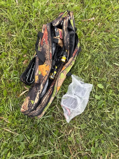 The image presents an outdoor scene in Айдарлы, Kazakhstan, dominated by fishing equipment laid out on natural ground. A multi-compartment soft-sided fishing rod case, patterned in a camouflage design featuring autumnal shades of green, brown, and orange, rests partially open on lush green grass.
In the leftmost open zippered compartment of the case, several silver-colored metal items, resembling fishing rod holders or specialized tackle with U-shaped hooks, are visible. The main central compartment reveals a partially visible package with a red and white label, displaying Cyrillic text including "КУРИЦЫ" (Chicken) and "ГРИ" (Grill), likely indicating a food item. Below this, a distinctive gold and black fishing reel with a white spool is prominent, attached to or positioned near a black fishing rod segment. Other zippered compartments are either closed or contain obscured items.
Adjacent to the camouflage case, a transparent, crinkled plastic bag contains several dark, rectangular objects, possibly fishing tackle boxes, with subtle red accents visible through the plastic.
The ground consists of varied green grass, interspersed with some broader-leaf weeds and occasional dried plant debris. The lighting is even, suggesting an overcast day or indirect sunlight, with no harsh shadows present. No individuals are depicted, and no specific actions or interactions are taking place.