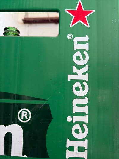 The image shows a close-up of a green Heineken beer crate. The crate features the iconic red star logo and the brand name "Heineken" printed vertically in white letters. A green glass beer bottle neck is visible in a cutout on the upper left side of the crate. The crate is made of plastic and shows some signs of wear and tear, including scuff marks and scratches. The background is out of focus, but appears to be a light-colored wall. The scene is outdoors, as indicated by the lighting and the presence of what appears to be a shelf in the background. There are no people visible in the image.