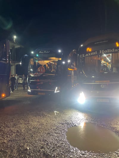 The image depicts a nighttime scene in Katauhar Kalan, India, featuring several parked buses. Two buses are prominent in the foreground. The bus on the right is a "Laxmi Holiday" bus, with "JAI MAA BAGLAMUKHI" written above the windshield and an "ALL INDIA TOURIST PERMIT" sticker. Its headlights are on, illuminating a large puddle of muddy water on the gravelly ground. The bus on the left has "JAI BAJARANGBALI" and "Ram Dalal" written on it. Several people are visible standing near the buses, some appearing to be passengers or staff. The environment is dark and appears to be an outdoor parking area or roadside, with some structures and lighting visible in the background. The ground is uneven, covered with gravel and dirt, and shows signs of recent rainfall with several puddles.